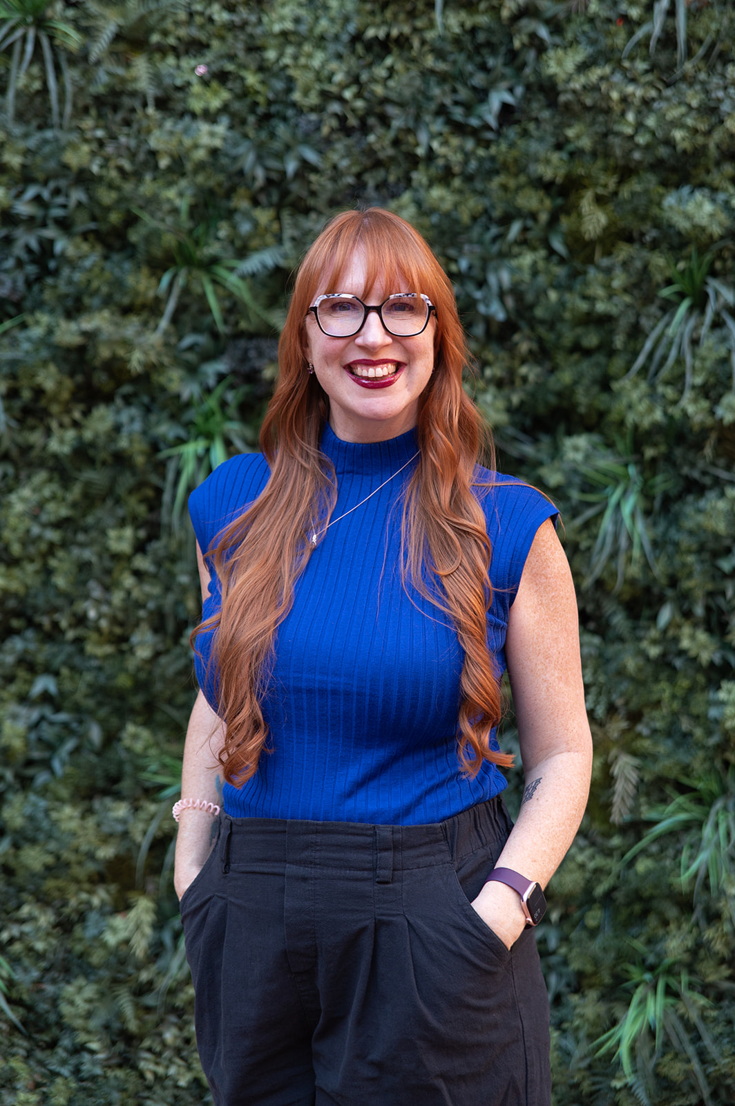 A woman with long red hair and glasses standing in front of a wall of a deep green plants